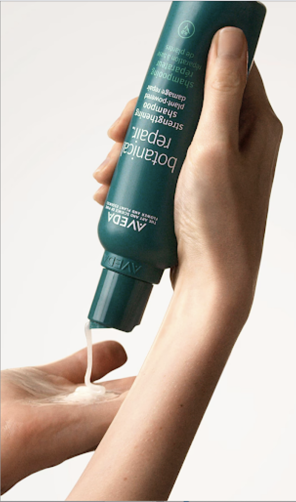 Hand squeezing a green bottle of Aveda Botanical Repair Strengthening Shampoo onto another hand. - Scott J Salons in New York, NY